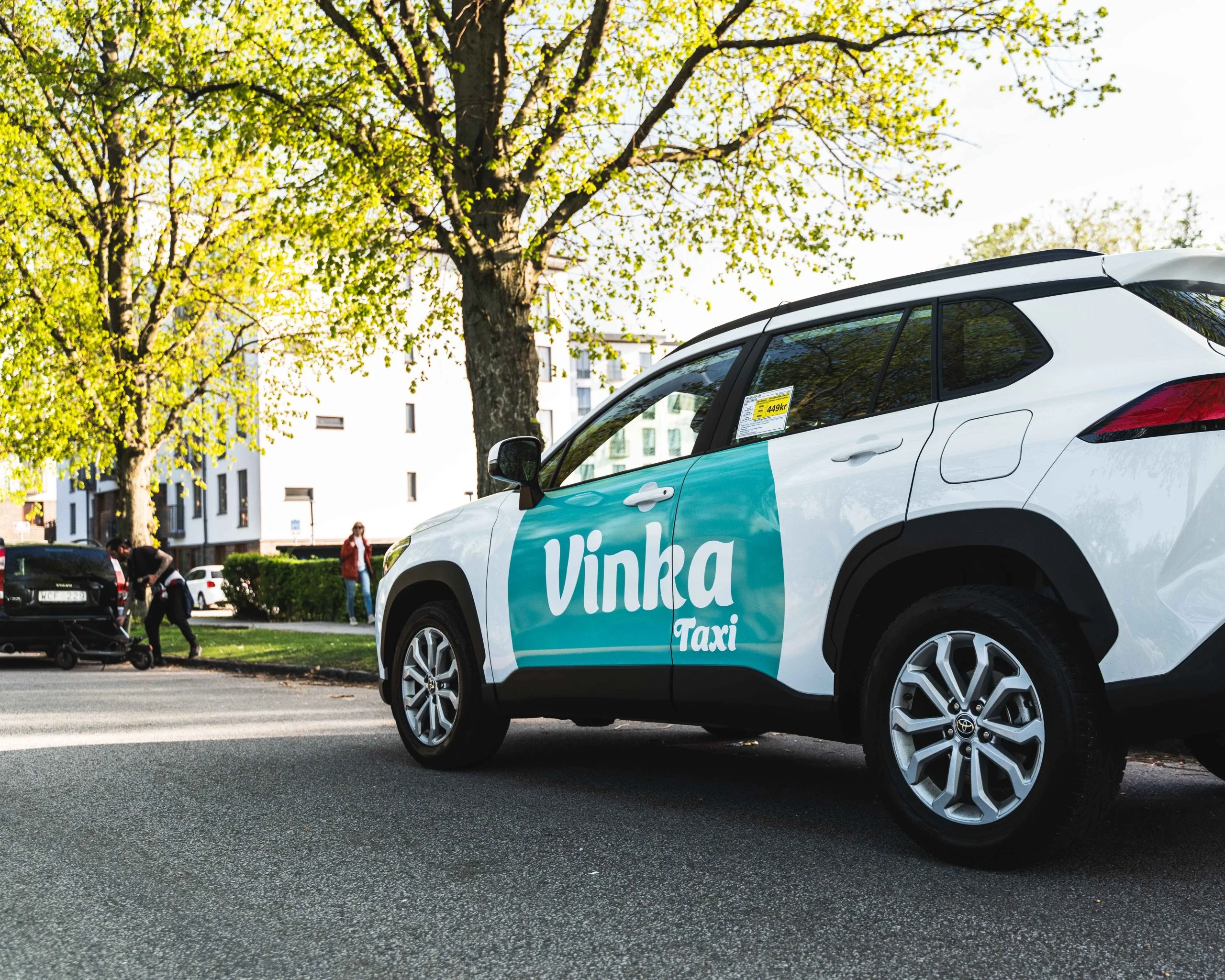 En vit Vinka Taxi med turkos logotyp på en solig, trädkantad gata